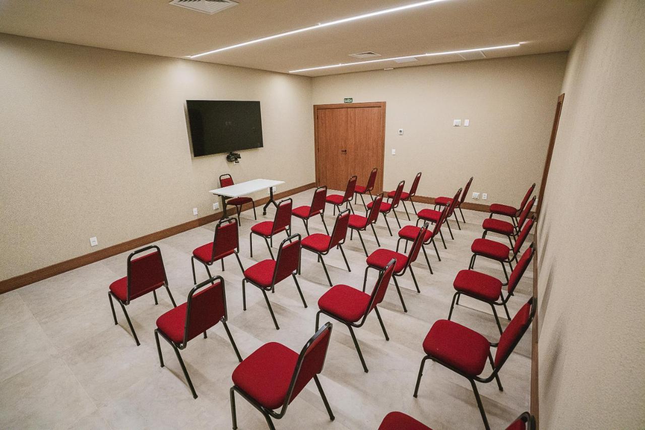 Buona Vitta Gramado Resort & Spa By Gramado Parks Kültér fotó The photo shows a conference or meeting room with a simple and modern design. There are several rows of red chairs arranged in a semi-circle facing a white table at the front. On the wall opposite the chairs, there is a flat-screen television mounted