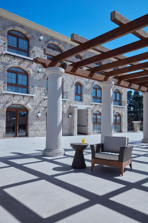 Buona Vitta Gramado Resort & Spa By Gramado Parks Kültér fotó The photo shows a well-designed outdoor space with a stone wall building in the background. There are several tall columns that support a wooden pergola overhead, creating a shaded area. In this space, there is a stylish chair and a small table besid