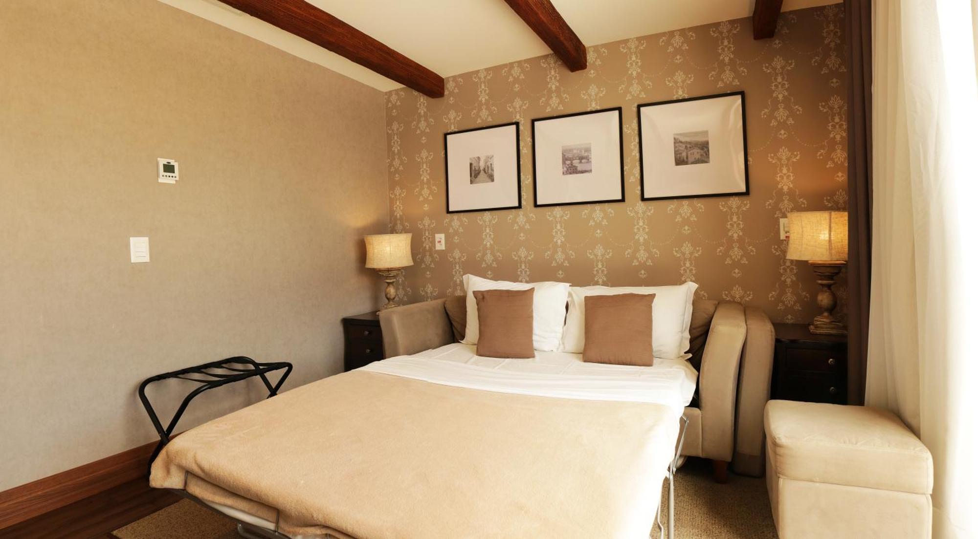 Buona Vitta Gramado Resort & Spa By Gramado Parks Kültér fotó The photo shows a cozy bedroom with a neutral color palette. There is a bed with white linens and two brown decorative pillows. On either side of the bed, there are bedside tables with lamps. The walls feature a subtle floral pattern, and three frame