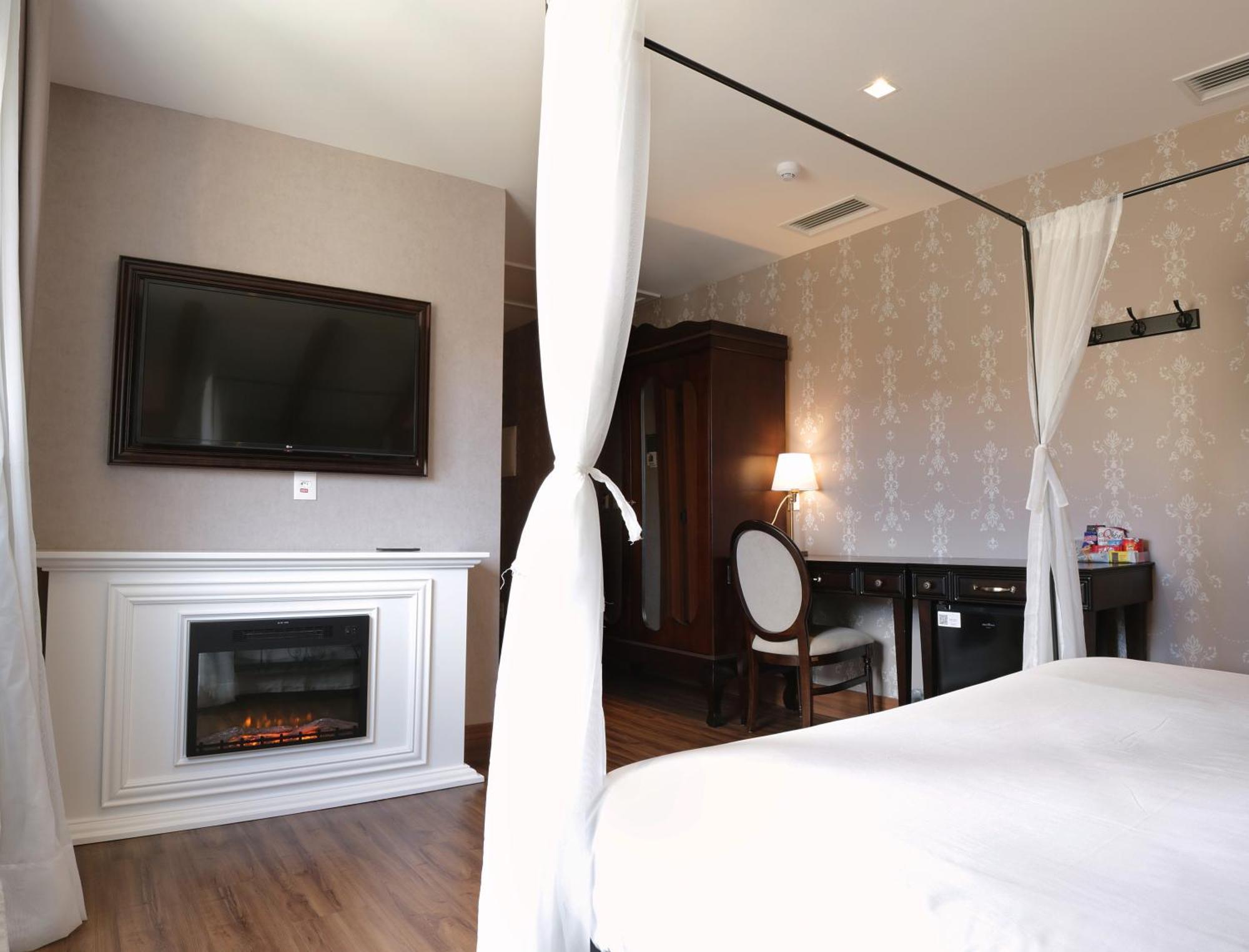 Buona Vitta Gramado Resort & Spa By Gramado Parks Kültér fotó The photo shows a cozy hotel room with elegant decor. There is a white bed with a canopy, and a modern television mounted on the wall across from it. To the side, a fireplace is built into a white mantel, adding a warm touch to the space. There's a w