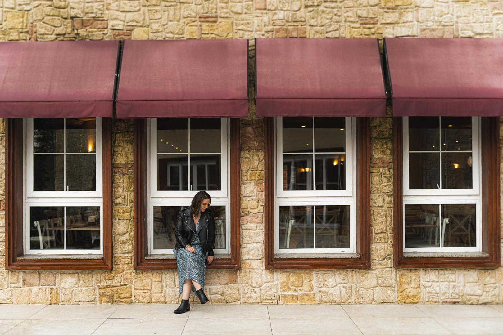 Buona Vitta Gramado Resort & Spa By Gramado Parks Kültér fotó The photo shows a woman standing against a stone wall with three large windows in front of her, each framed with wooden borders. The windows have maroon awnings above them. The woman is dressed in a long skirt and leather jacket, leaning slightly aga